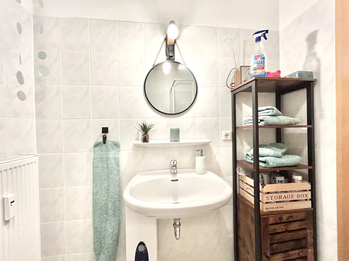 ferienwohnung-cottbus-green-apartment-badezimmer Modernes Waschbecken mit rundem Spiegel und Holzregal im Badezimmer des Green Apartments – Ihre gepflegte Ferienwohnung in Cottbus.